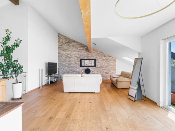 Séjour lumineux sous combles avec parquet en chêne, canapé clair et mur en pierre naturelle à Chernex