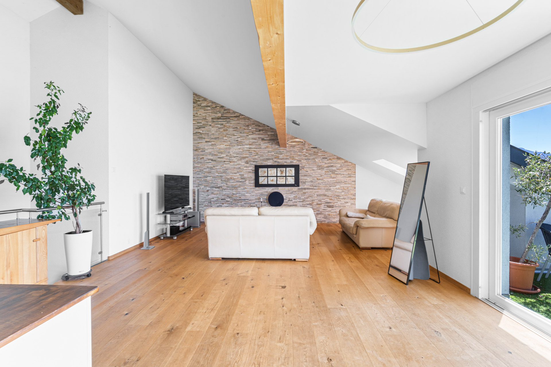 Séjour lumineux sous combles avec parquet en chêne, canapé clair et mur en pierre naturelle à Chernex