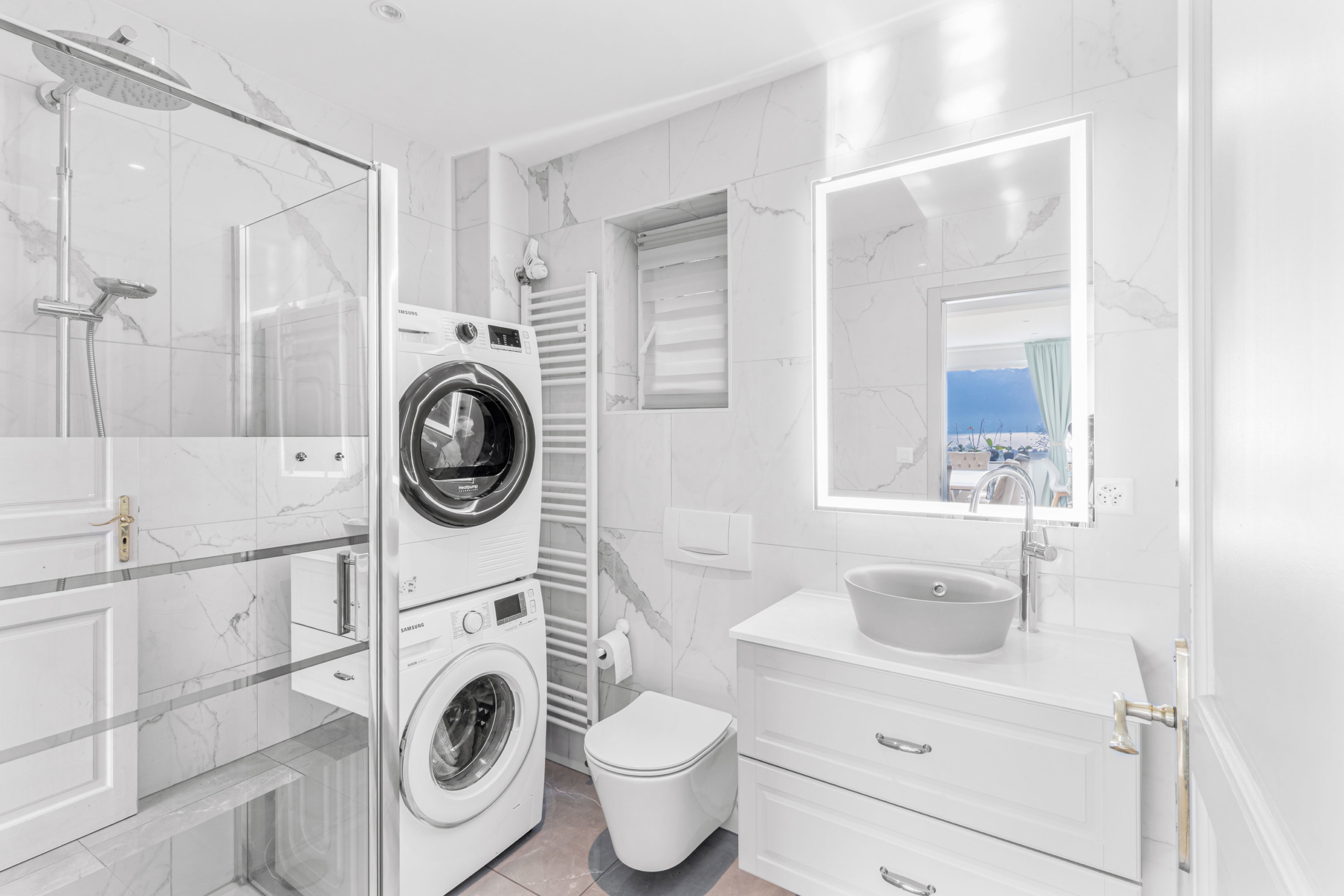 Salle de bain moderne avec douche italienne vitrée, machine à laver et sèche-linge intégrés, vasque design et miroir lumineux.