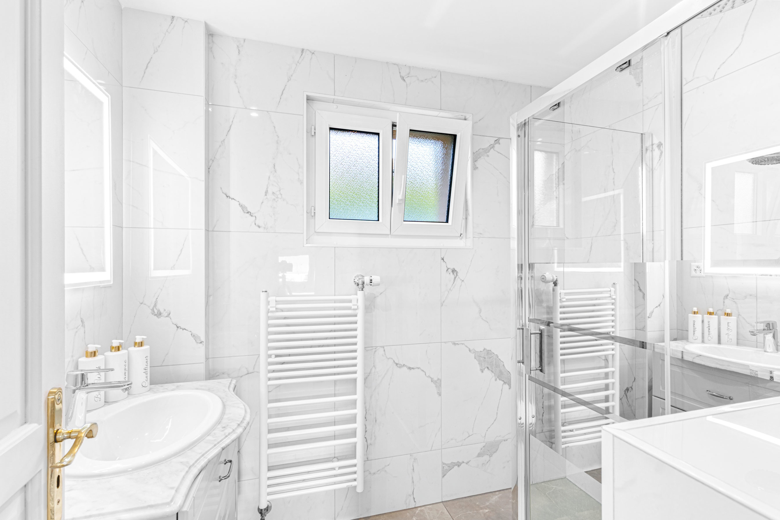Salle de bain avec douche vitrée, meuble vasque en marbre et fenêtre à double battant.