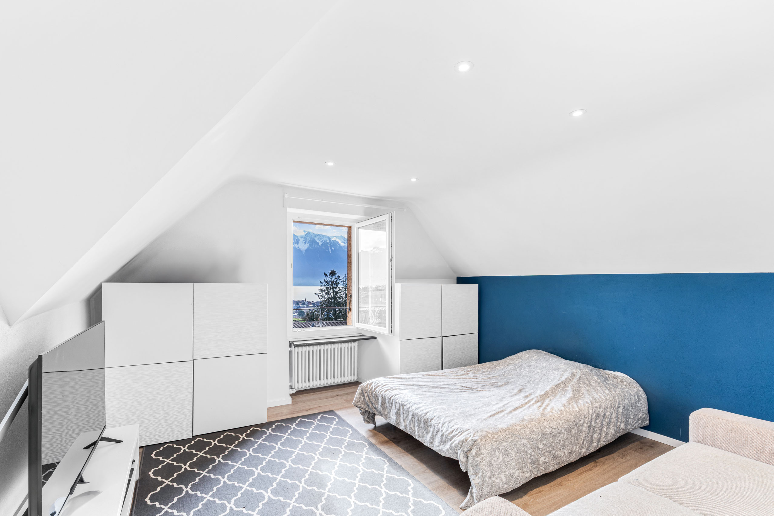 Chambre mansardée avec lit double, murs blancs et mur accent bleu, vue sur les Alpes.