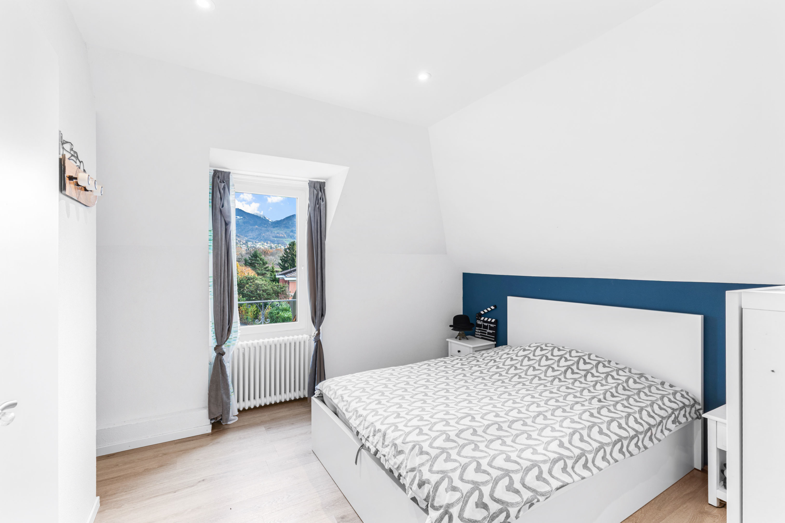 Chambre d’enfant ou d’amis avec lit double, radiateur ancien et fenêtre avec rideaux gris.