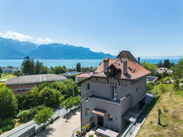 Façade de la maison avec vue sur le lac Léman, montagnes en arrière-plan et ciel dégagé.