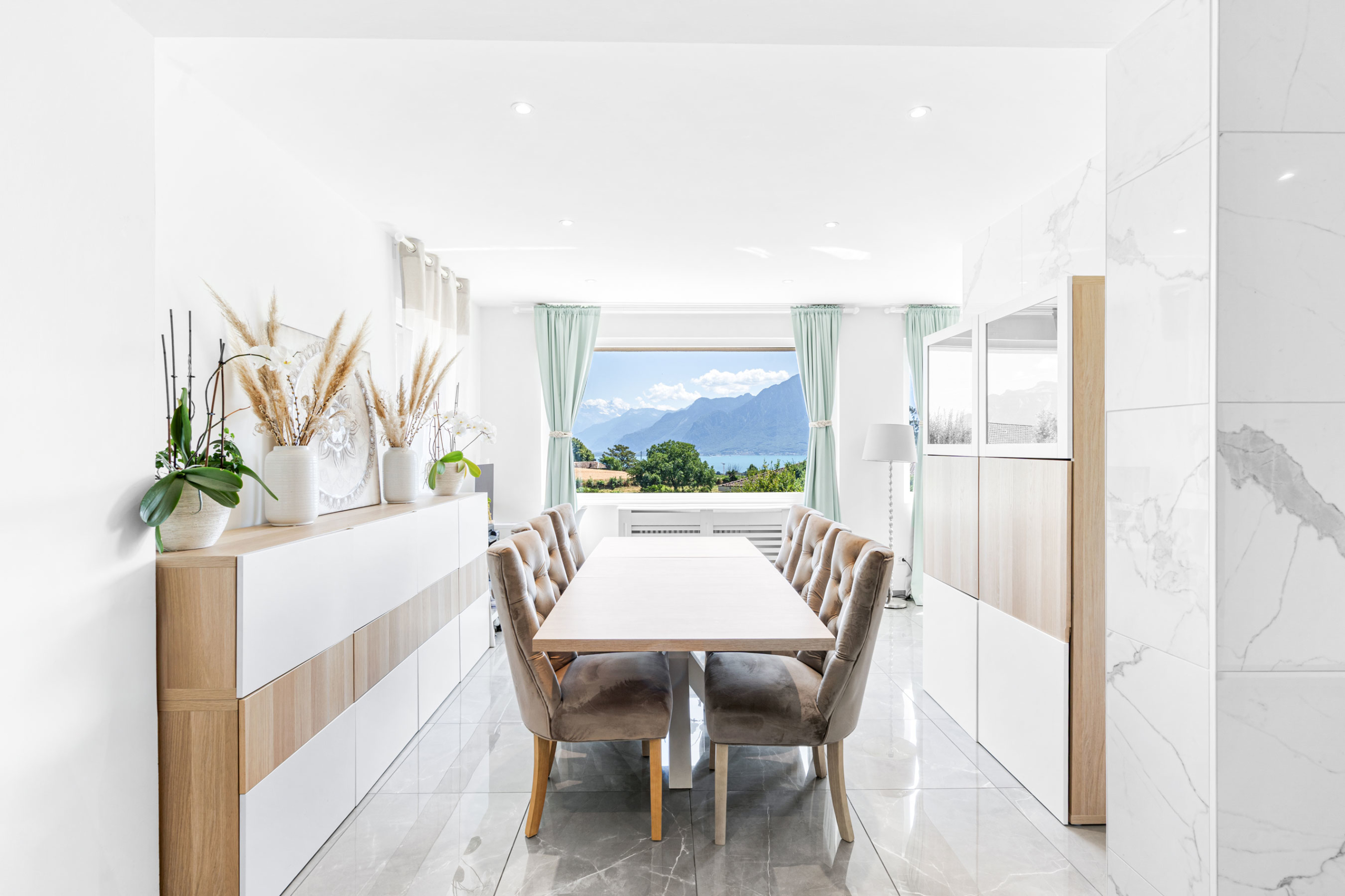 Salle à manger avec grande table en bois, chaises capitonnées et vue sur les montagnes.