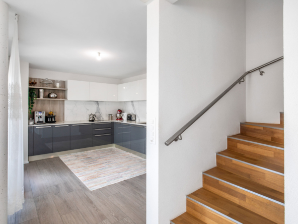 Vue sur une cuisine contemporaine avec meubles laqués gris et blancs, et escalier en bois menant à l’étage d’une villa à Jongny.