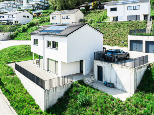 Maison moderne avec grande terrasse, panneaux solaires sur le toit, place de parc extérieure et vue sur le quartier résidentiel de Jongny.