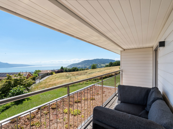 Balcon couvert avec canapé d’extérieur, vue panoramique sur les champs, les toits du quartier et le lac Léman.