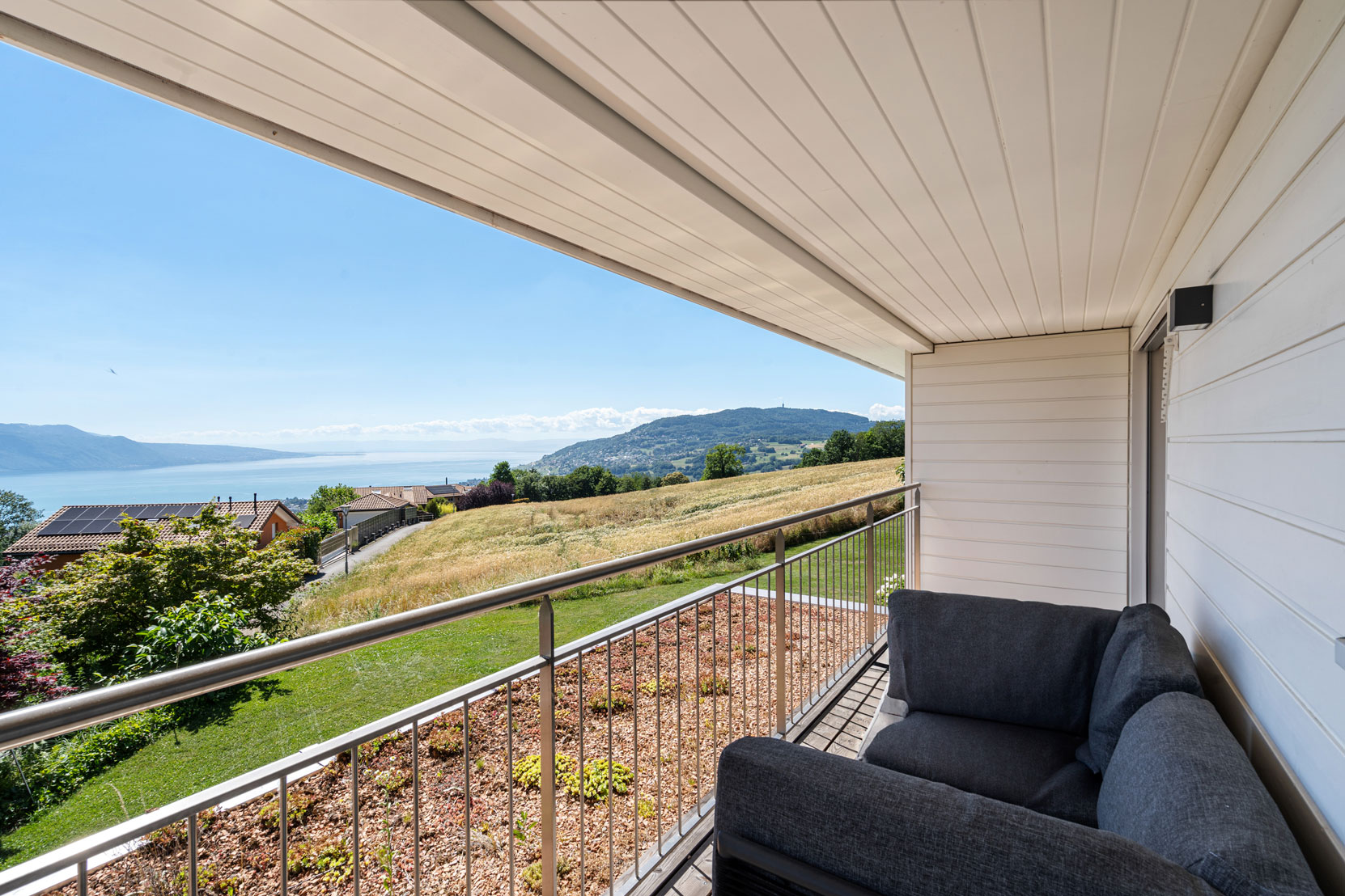 Balcon couvert avec canapé d’extérieur, vue panoramique sur les champs, les toits du quartier et le lac Léman.