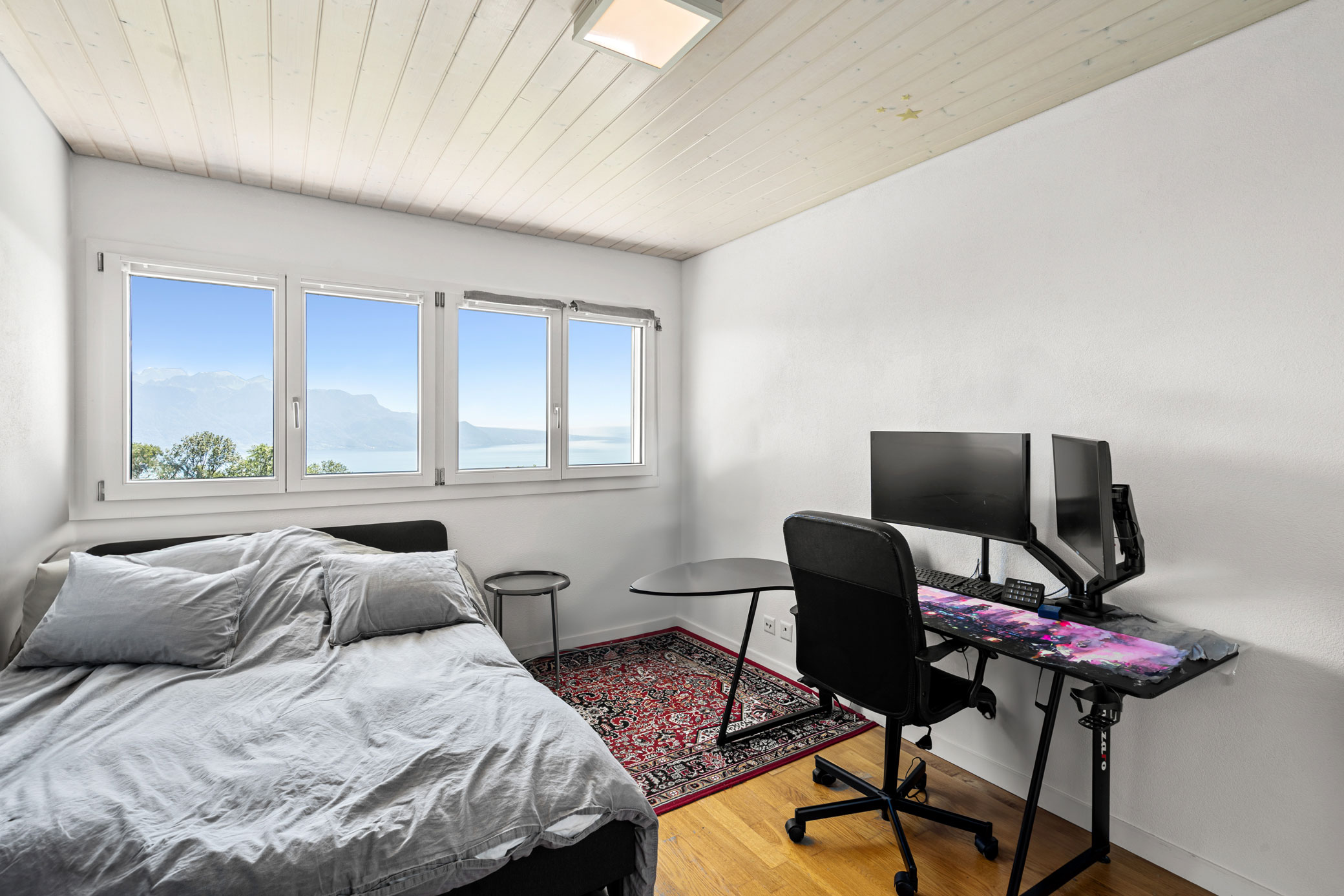 Chambre équipée d’un lit double et d’un bureau informatique, avec grande fenêtre et vue sur le lac Léman.