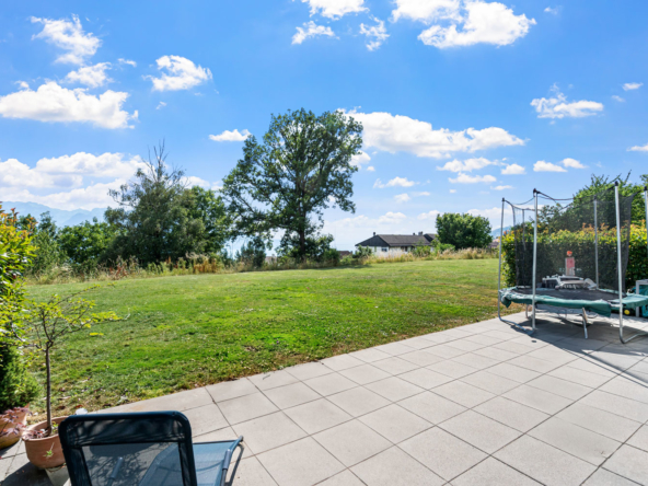 Vue dégagée sur une pelouse arborée depuis une terrasse avec mobilier et trampoline