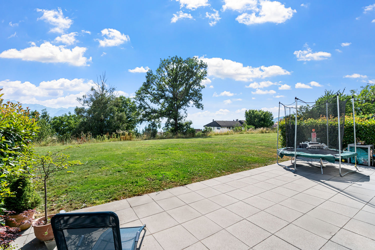 Vue dégagée sur une pelouse arborée depuis une terrasse avec mobilier et trampoline