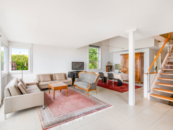 Salon lumineux avec mobilier classique, grande baie vitrée et escalier en bois menant à l’étage.