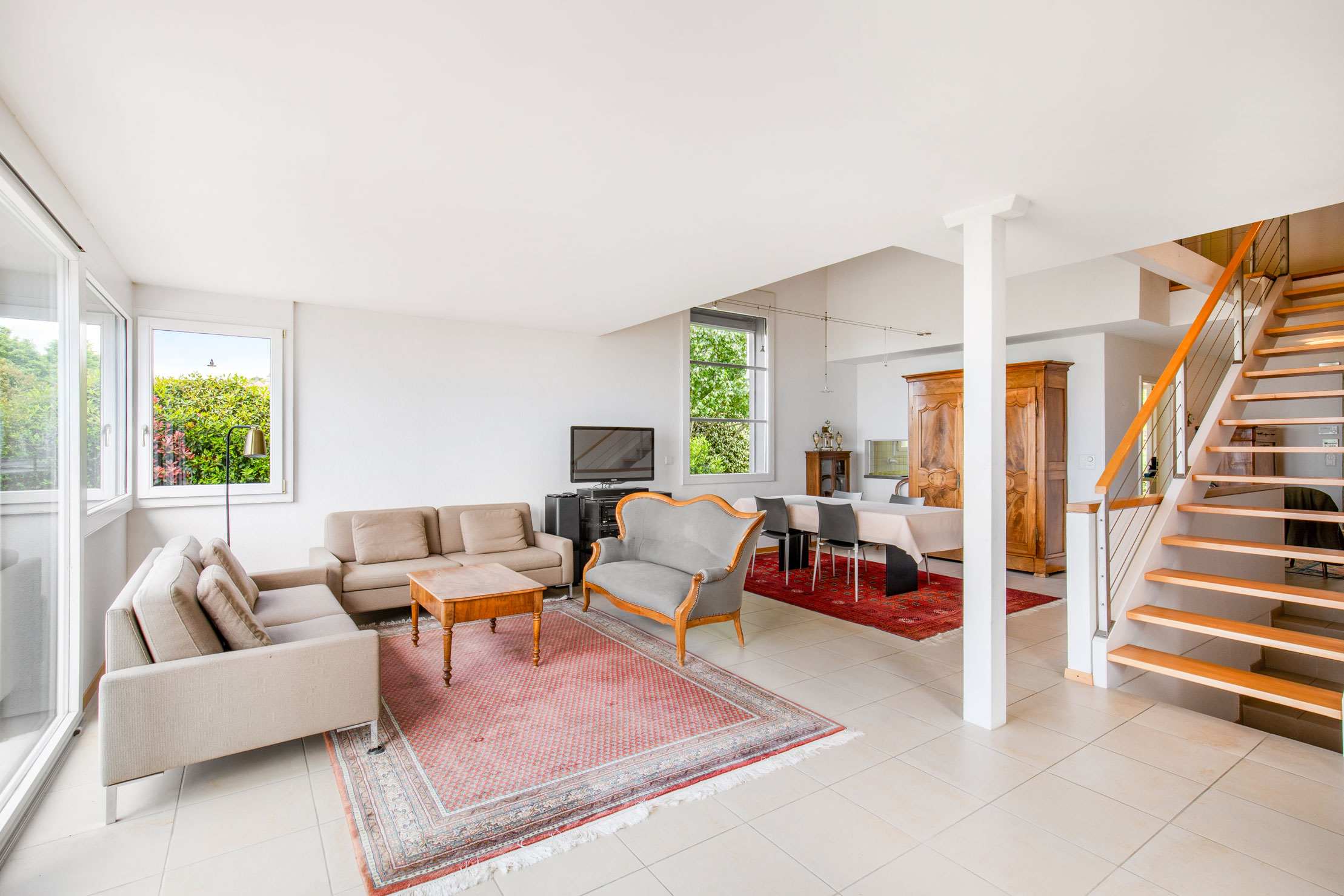Salon lumineux avec mobilier classique, grande baie vitrée et escalier en bois menant à l’étage.