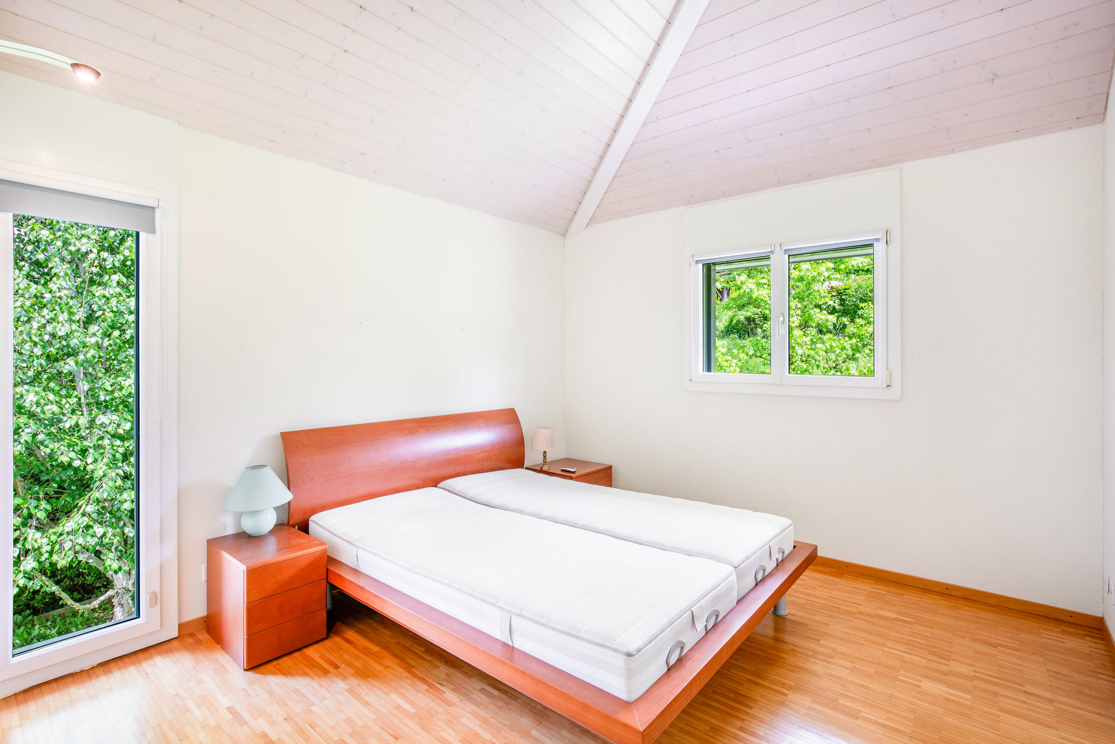Chambre avec lit double en bois, plafond en pente et vue sur jardin arboré.