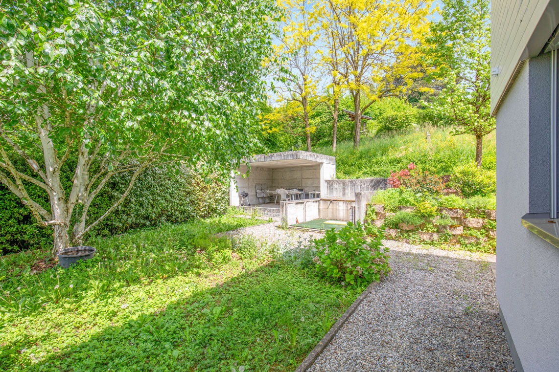 Jardin arboré avec coin repas couvert en béton, cadre végétal naturel.