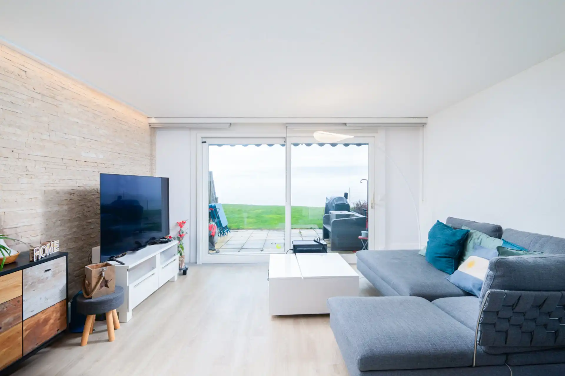 Salon lumineux avec canapé gris et grande baie vitrée donnant sur terrasse et jardin