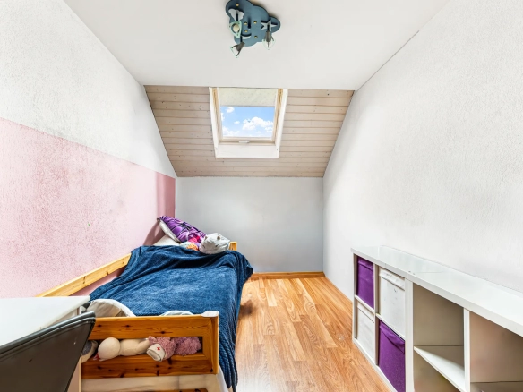 Petite chambre mansardée lumineuse avec un lit simple, du parquet et une fenêtre de toit.