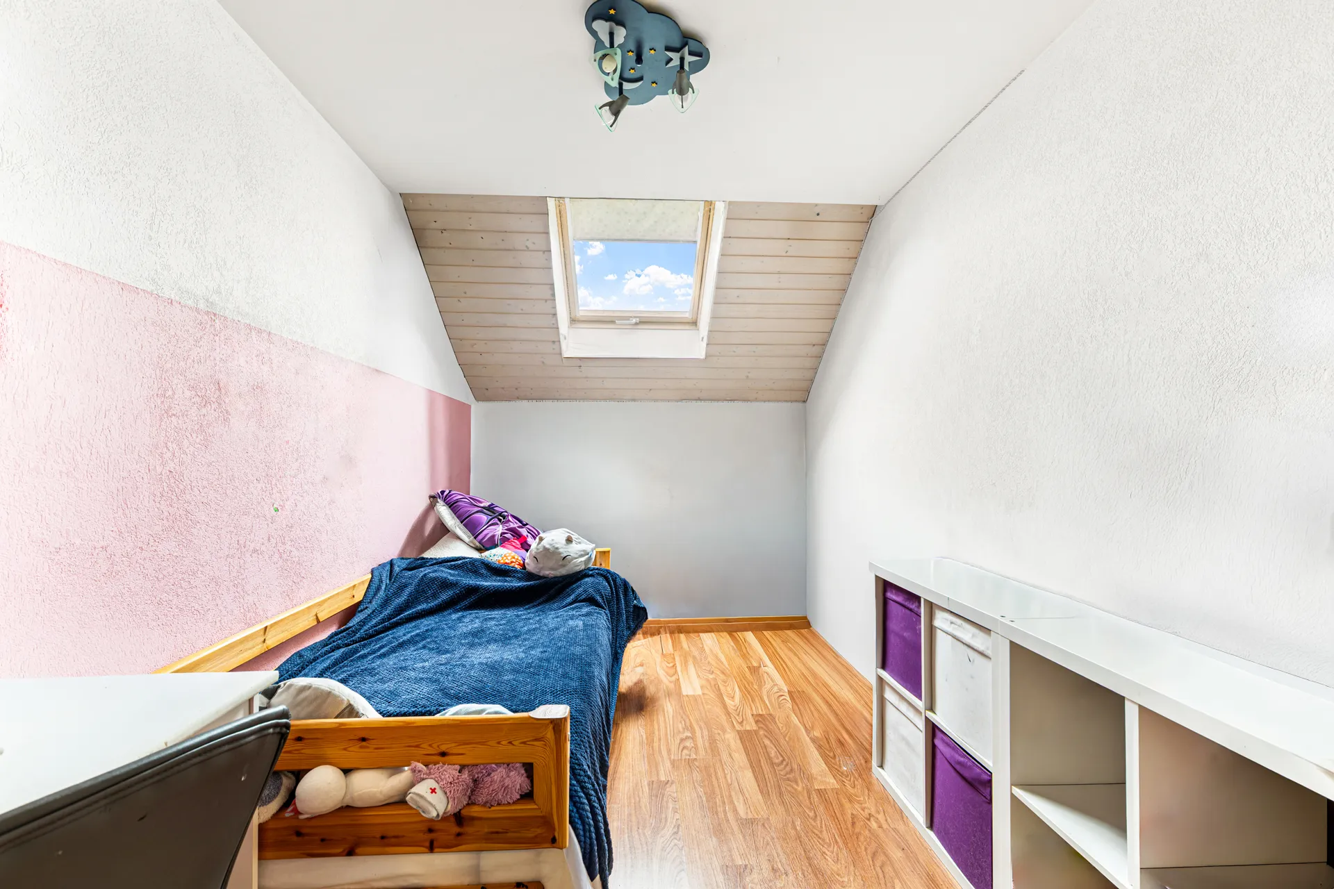 Petite chambre mansardée lumineuse avec un lit simple, du parquet et une fenêtre de toit.
